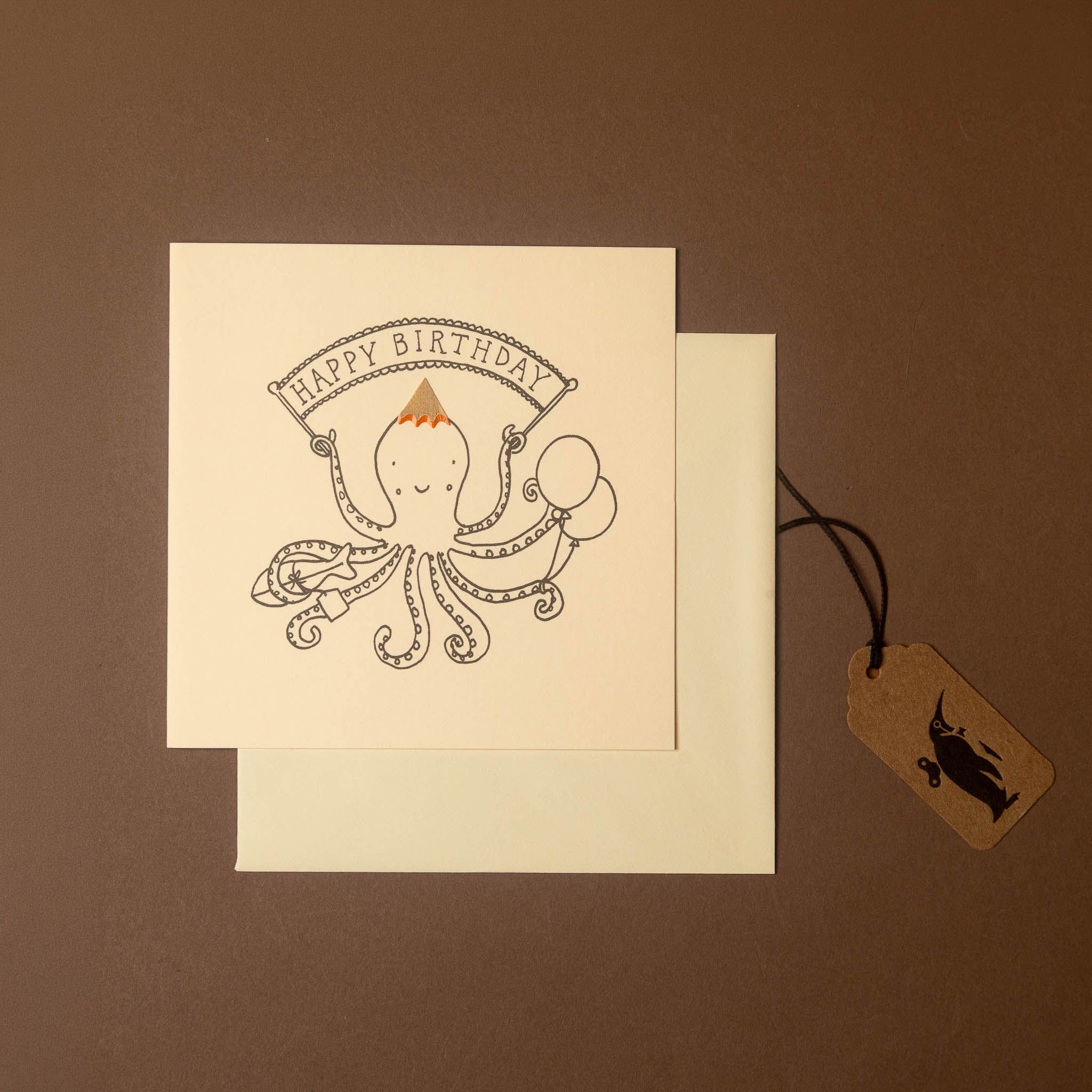 A square birthday card with a black line-art octopus illustration and a banner reading “HAPPY BIRTHDAY,” shown with a cream envelope and a small kraft gift tag on a brown background.