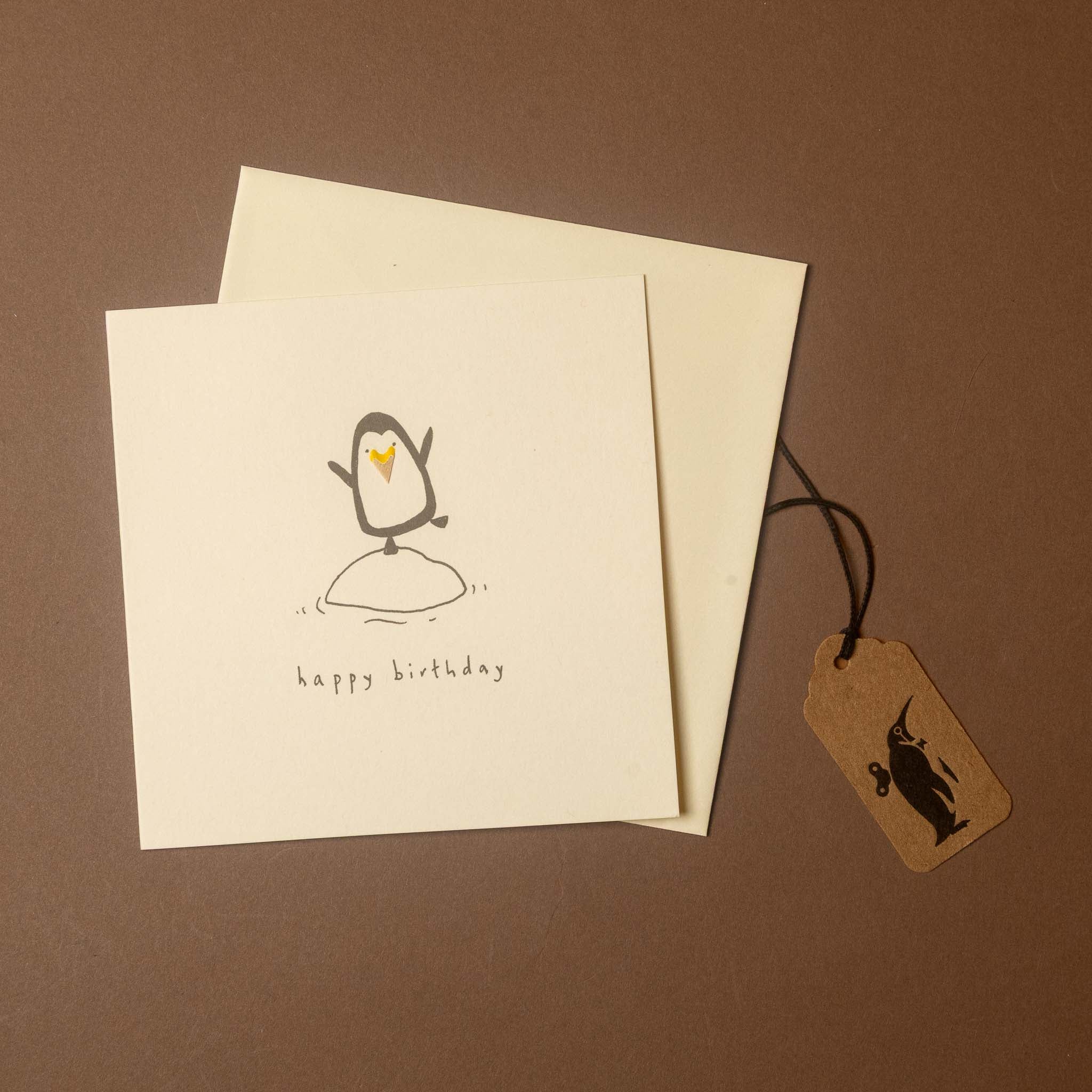 A square cream birthday card with a simple penguin drawing and the words 'happy birthday' is shown on a brown background, with a matching cream envelope behind it and a small kraft paper tag attached by a black string.