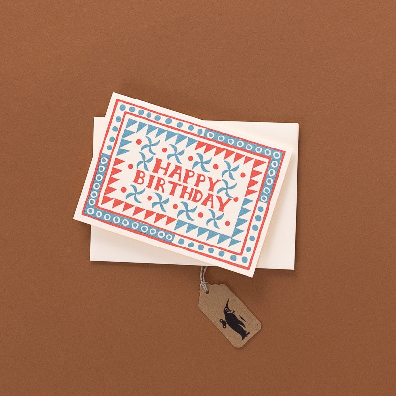 A square greeting card with a red and blue geometric border and the words 'HAPPY BIRTHDAY' in red, placed on top of a white envelope against a brown background. A small kraft paper tag hangs from the envelope and has a black cat silhouette printed on it.