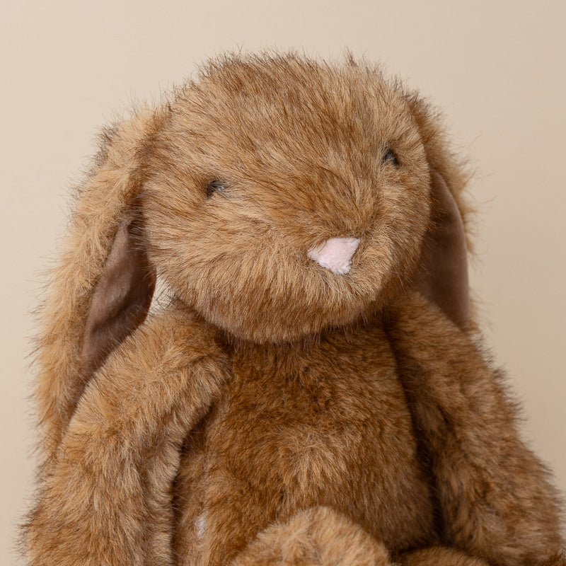 A close-up of a brown plush bunny toy with long floppy ears, a pale pink nose, and dark embroidered eyes, set against a plain beige background.