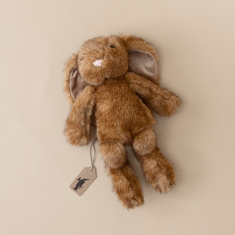 A plush brown bunny toy with long ears, a small white nose, and a hanging paper tag, photographed against a plain warm beige background.