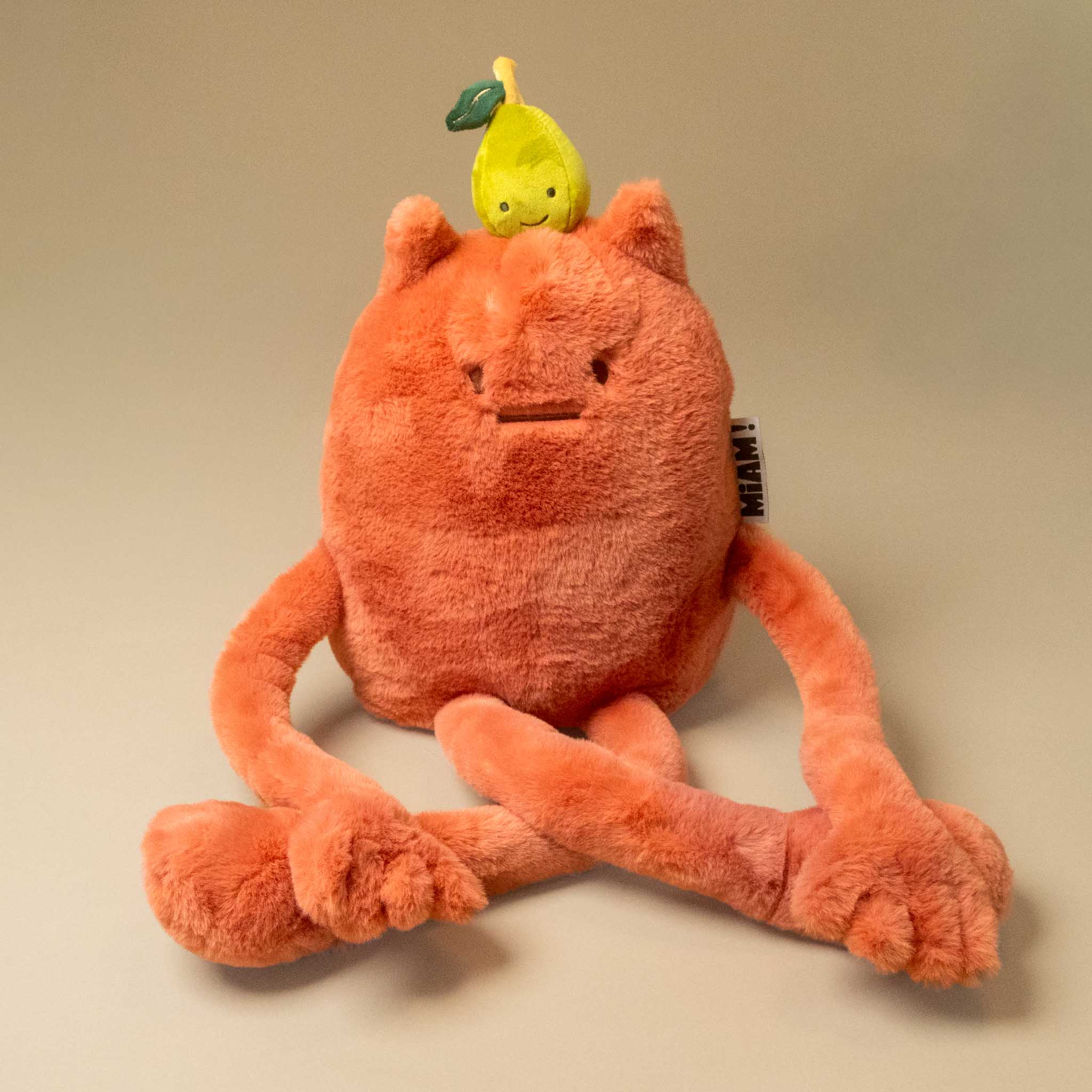 An orange plush britney-pear-grand toy with long limbs and a small yellow pear character on its head, photographed on a plain beige studio background.