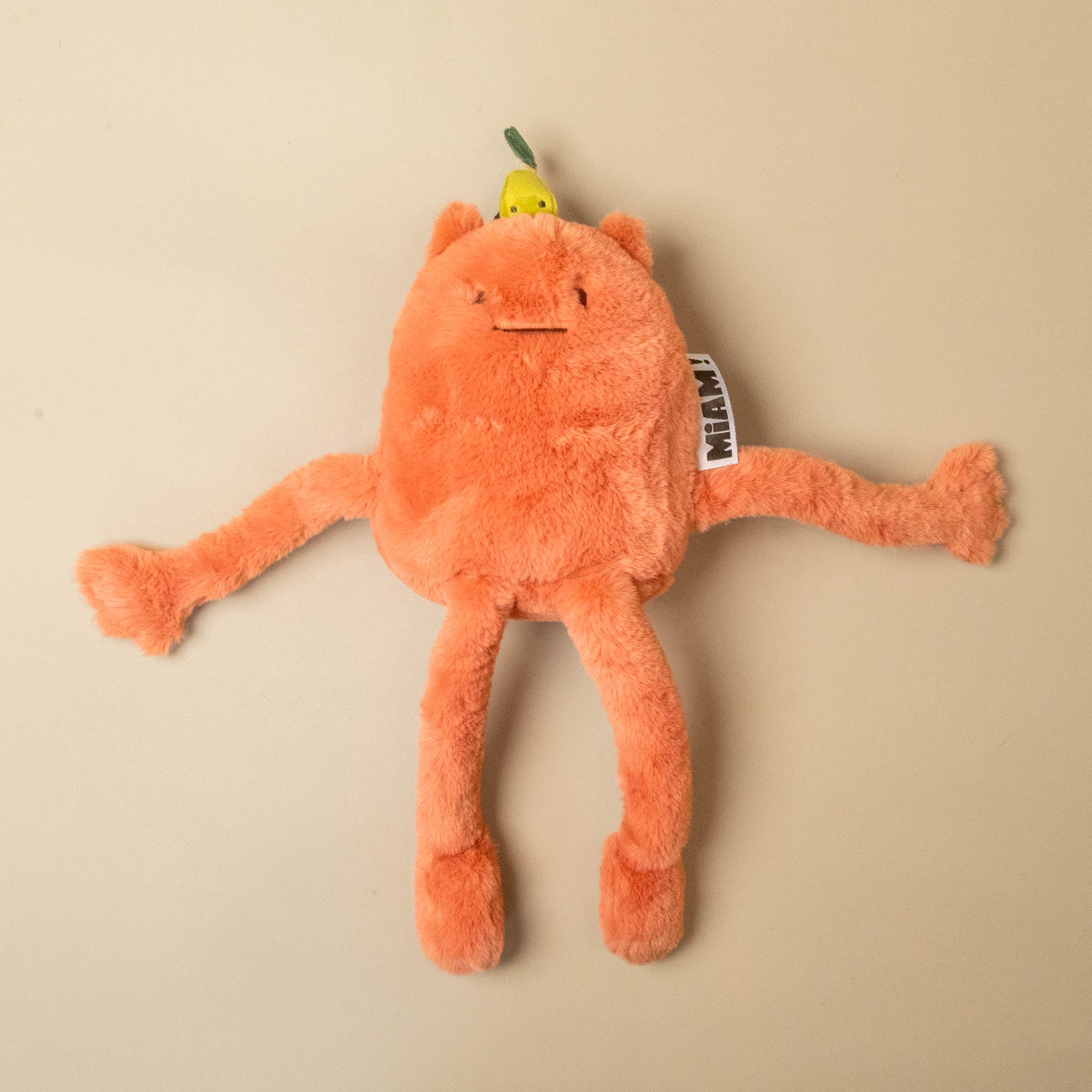 An orange plush britney-pear-petit toy with a small yellow pear on its head, photographed on a plain beige background