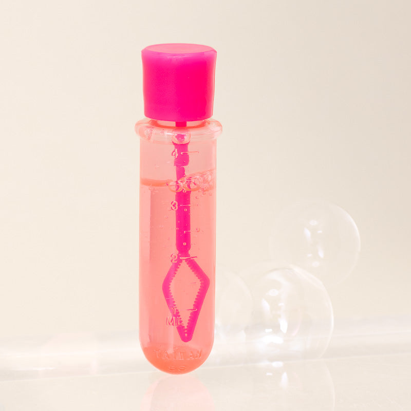 A clear plastic bubble wand bottle with a bright pink cap and internal bubble wand, filled with pink liquid, shown on a light beige background with soft blurred bubbles.