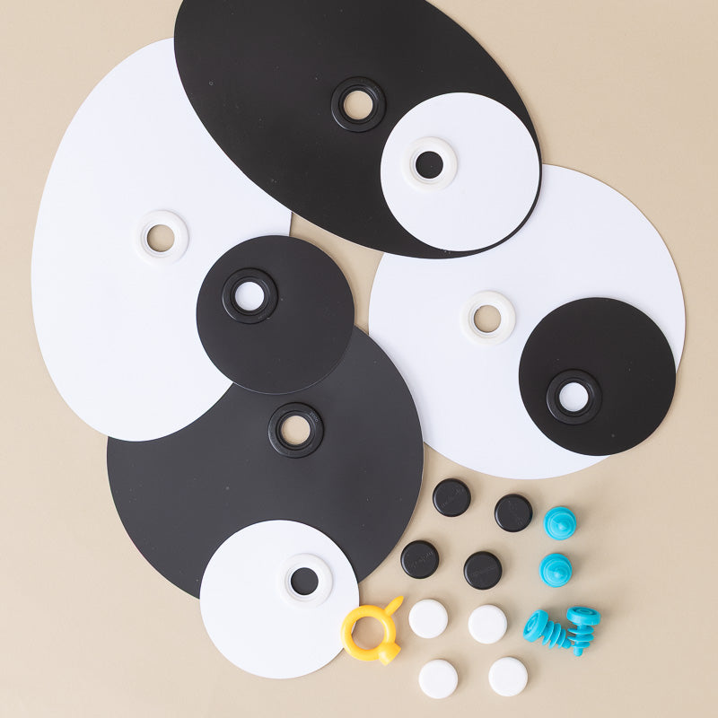 A set of black and white circular and oval pieces arranged on a beige background, with small colored studs and a yellow-and-blue hook-like accessory nearby.
