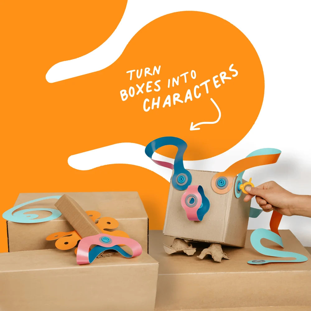 A hand is assembling a cardboard box character toy with colorful paper wings and buttons, set against a bright orange and white background with the text 'TURN BOXES INTO CHARACTERS'.