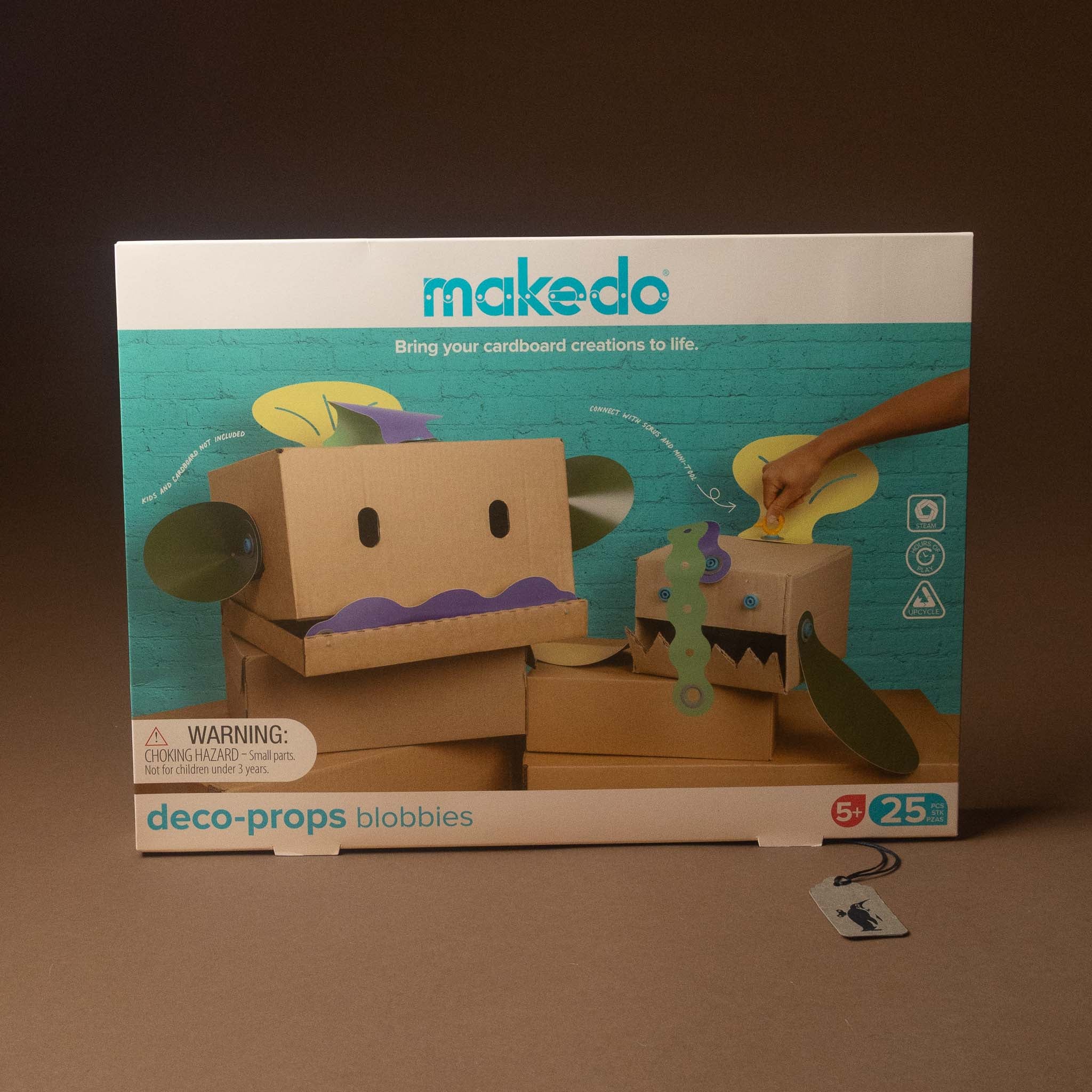 Makedo Deco-Props Blobbies toy kit box, showing cardboard creature figures and a hand interacting with the packaging, set against a dark brown background.