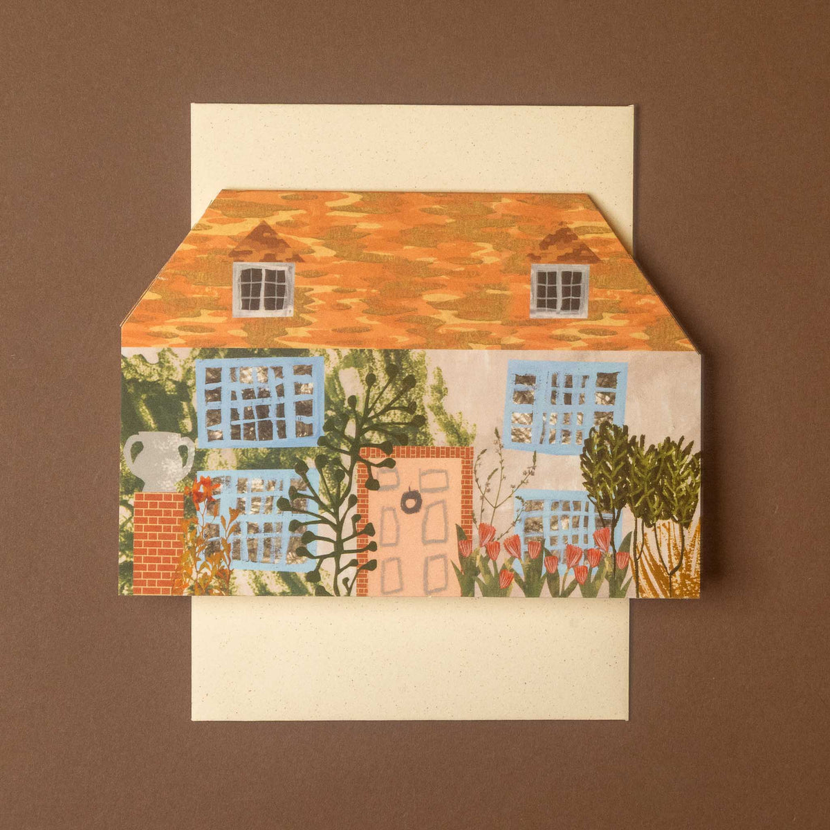 A square art print or card featuring a whimsical house illustration with an orange patterned roof, blue windows, green foliage, and a cream background, displayed on top of a cream envelope against a brown backdrop.