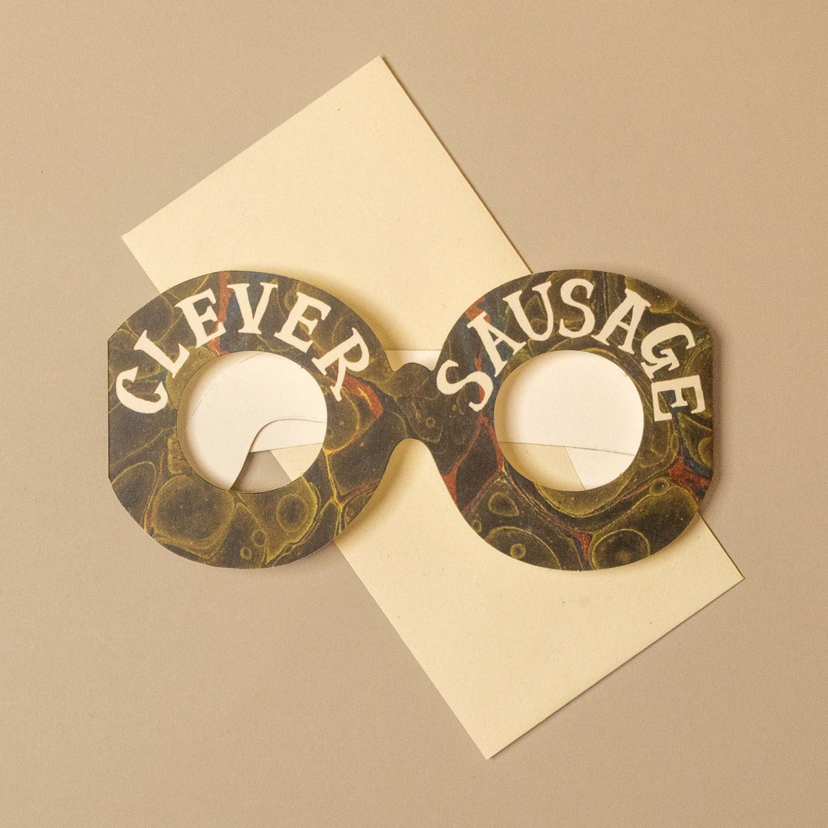 A novelty pair of round novelty glasses with a marbled pattern in dark green, gold, and black, featuring the words 'CLEVER SAUSAGE' printed in white on the front. The glasses are styled on top of a cream-colored envelope against a solid beige background.