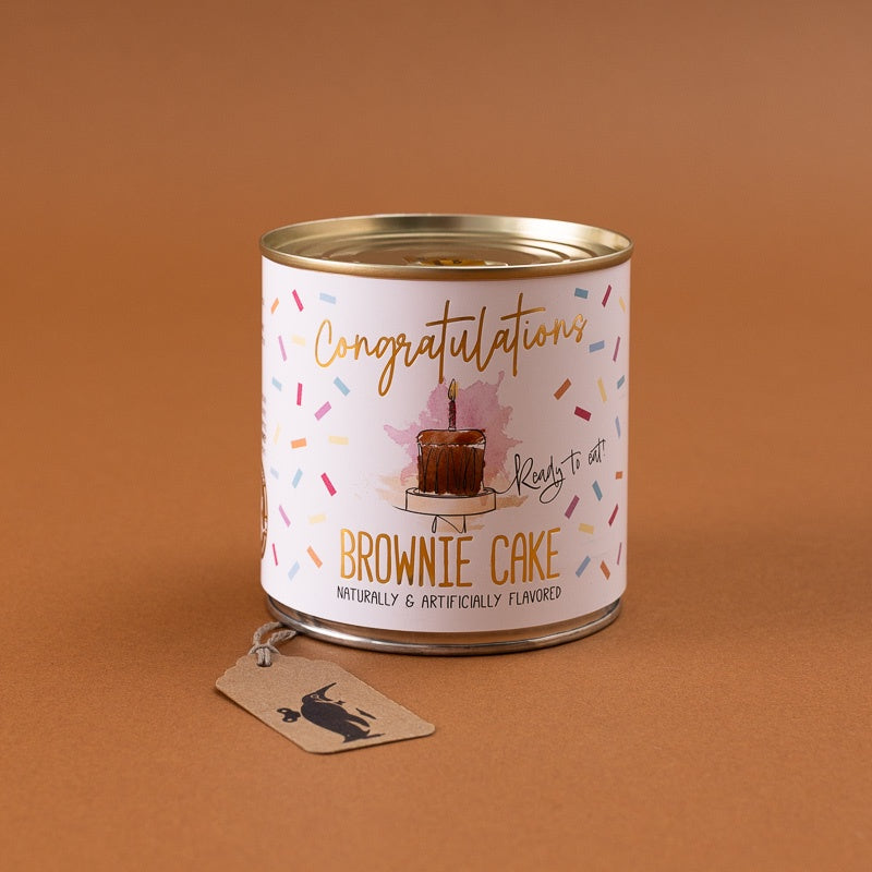A can of 'Congratulations Brownnie Cake' treats on a warm brown background, featuring a white label with gold and multicolor text and a small dangling paper tag.