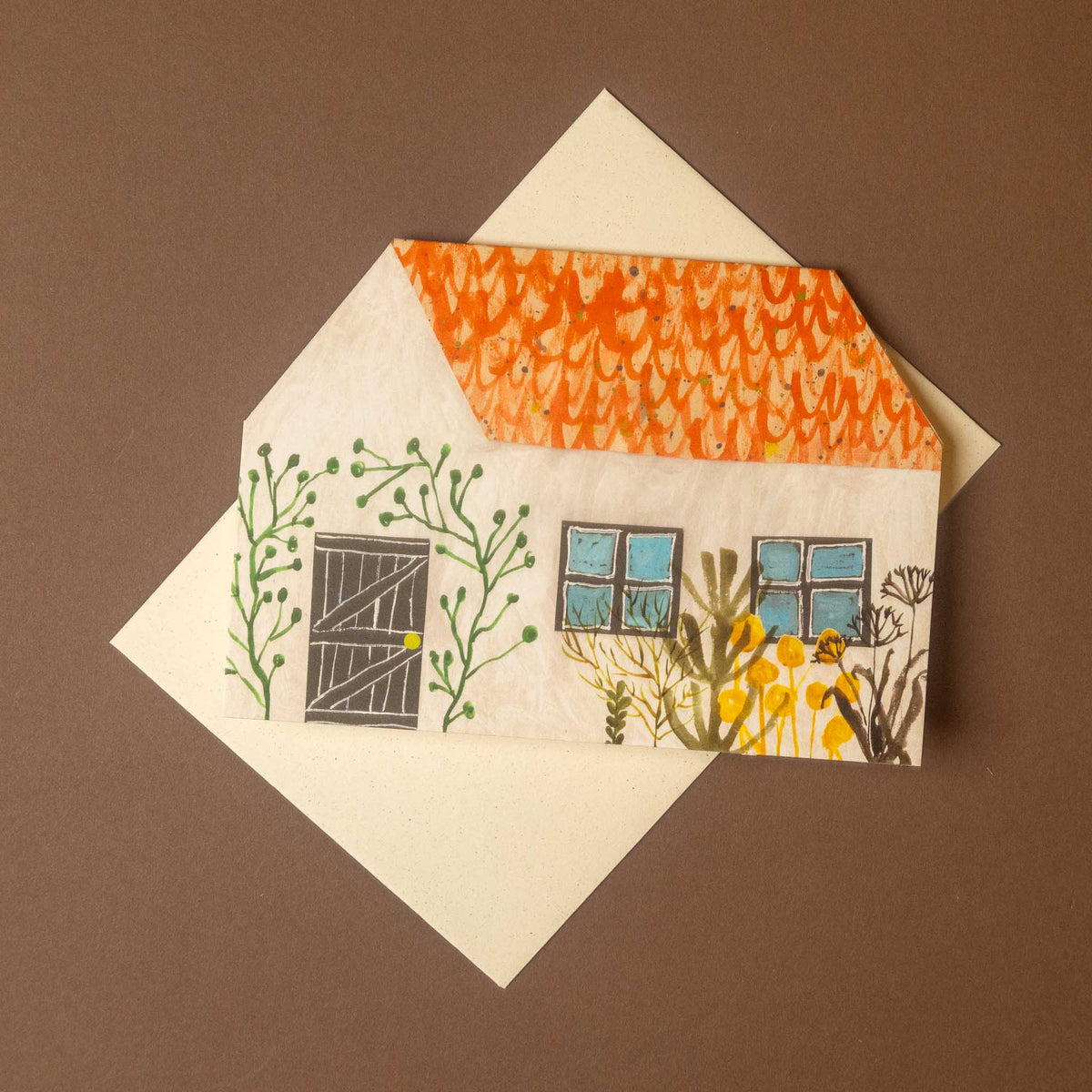 A square cream-colored greeting card featuring a hand-drawn illustration of a small house with an orange patterned roof, blue windows, green vines, and yellow flowers, shown with a matching cream envelope on a solid brown background.