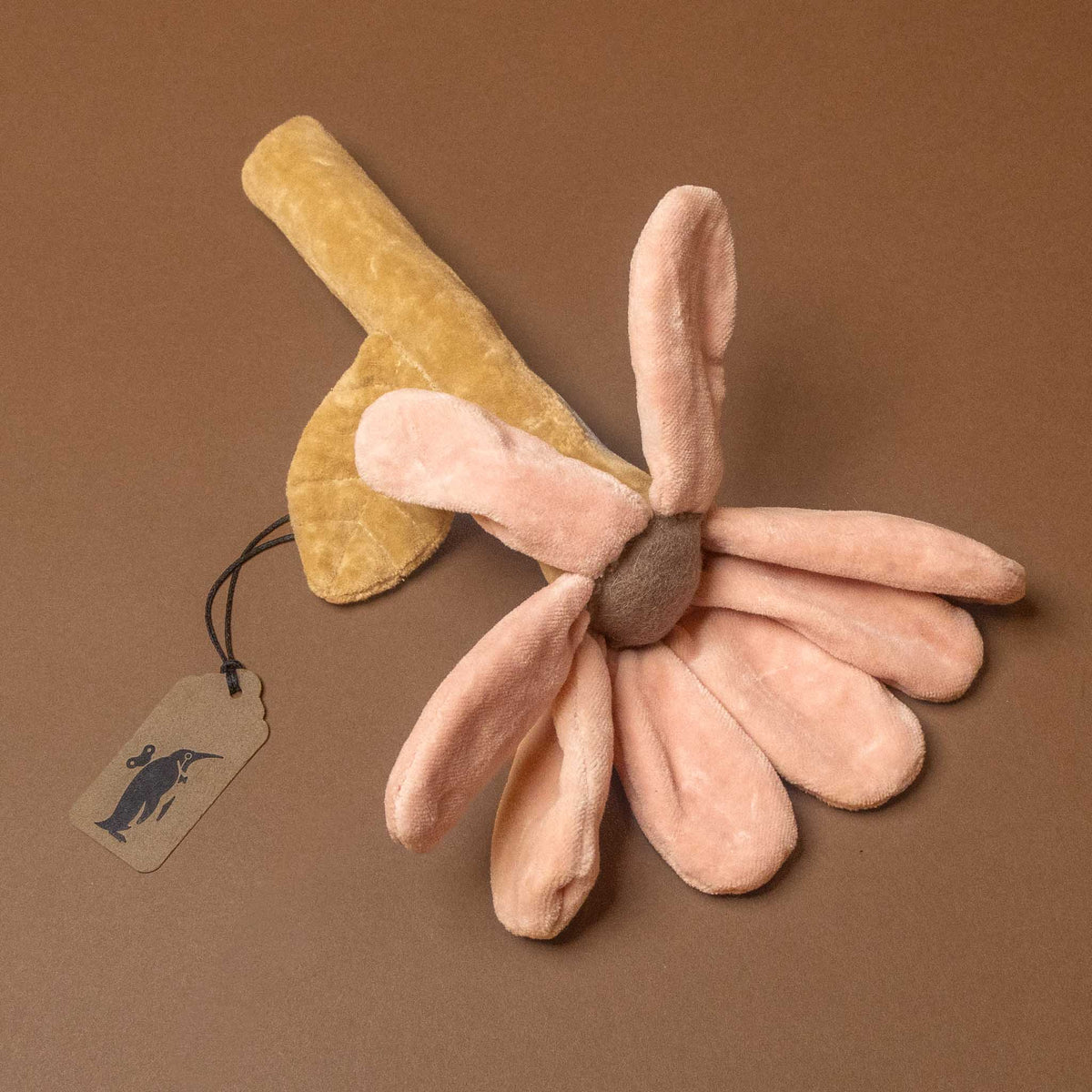 A plush flower-shaped cotton-flower-rattle in soft pink and tan, with a small paper hangtag attached, photographed on a solid brown background.