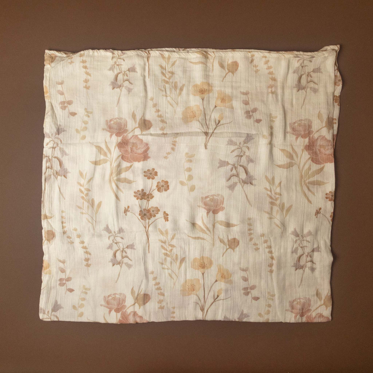 A square cotton muslin cloth with a delicate floral and botanical print in muted shades of pink, beige, and brown on an off-white background, laid flat against a solid dark brown backdrop.