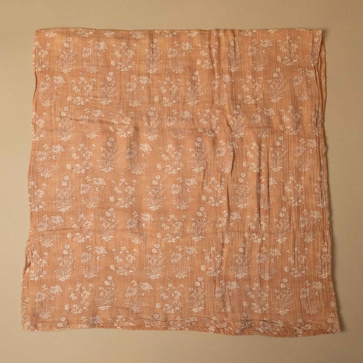 A square peach-colored muslin cloth with a white floral and botanical print, laid flat against a neutral beige background.
