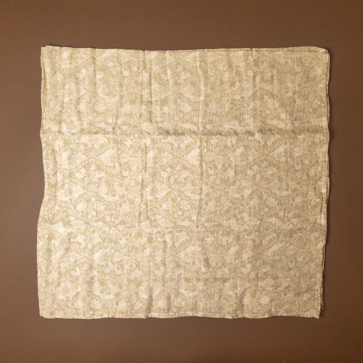 A square beige fabric cloth with a light brown damask pattern, laid flat against a solid brown background.