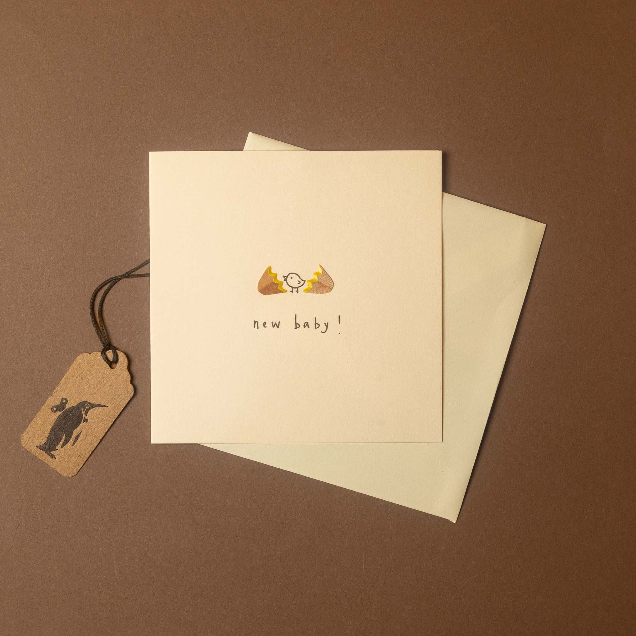 A square cream greeting card with a small hand-drawn baby illustration and the words 'new baby!' on the front, shown with a matching envelope and a small kraft paper tag on a brown background.