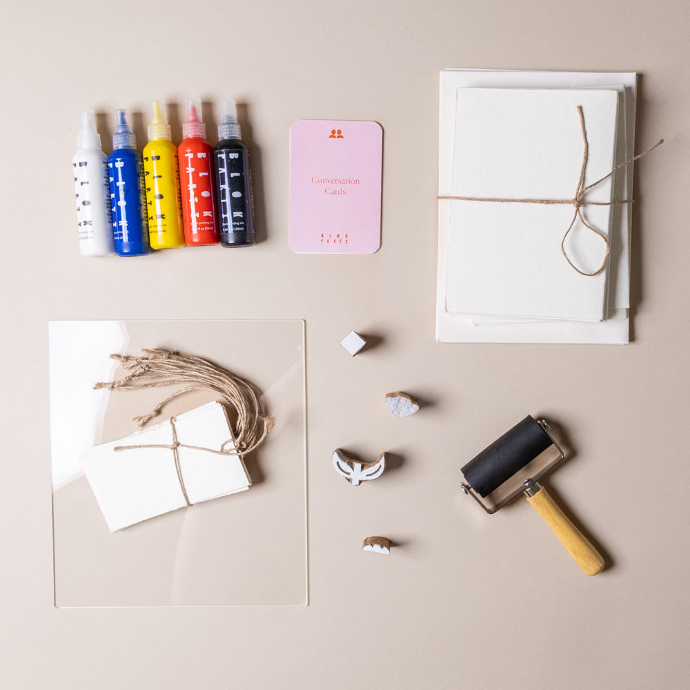 create-your-own-block-print-stationary-kit-tulip Colorful ink bottles, a pink card, white paper, and a roller on a beige surface