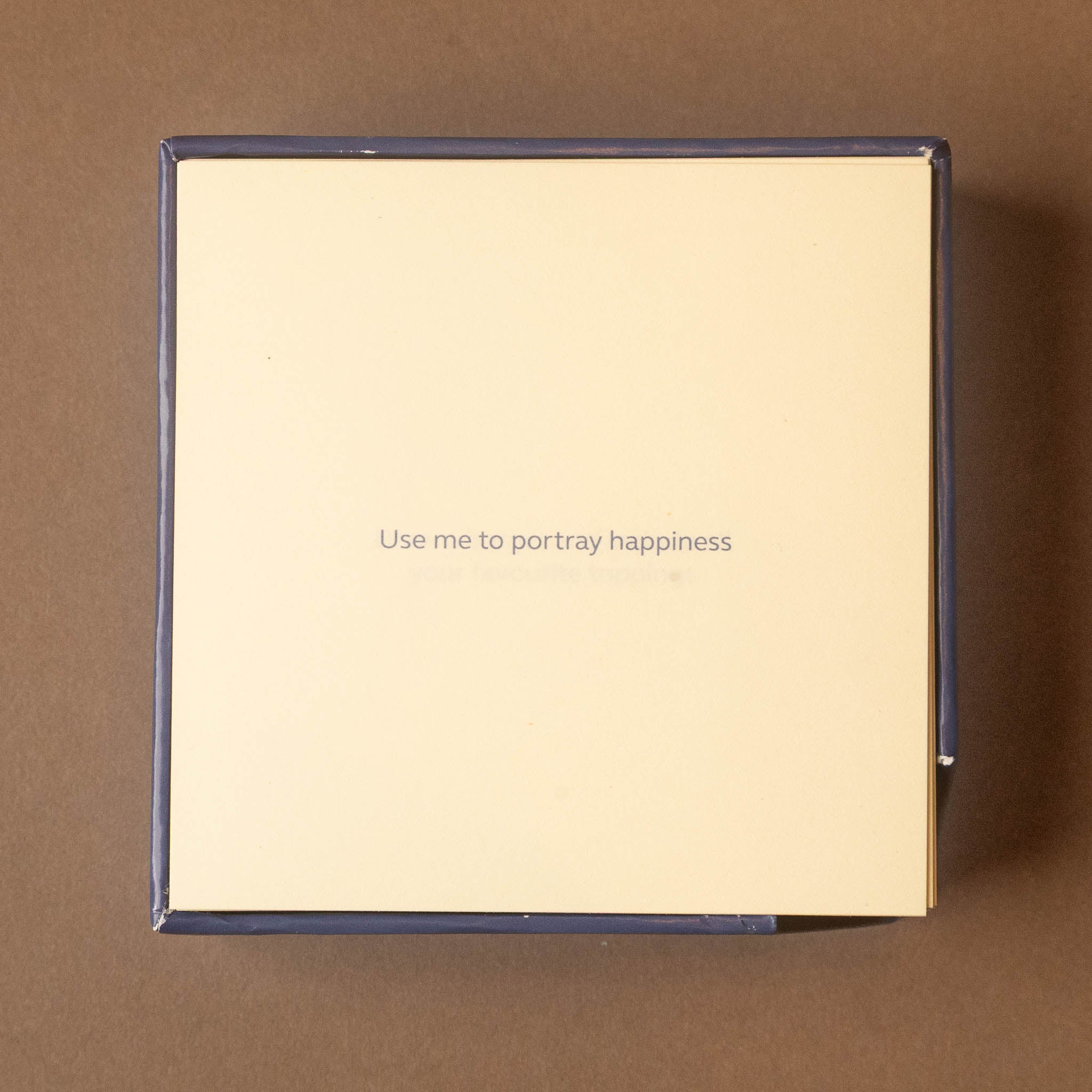 A square cream-colored product box with a dark navy blue border, featuring the printed text 'Use me to portray happiness' in the center, set against a solid brown background.