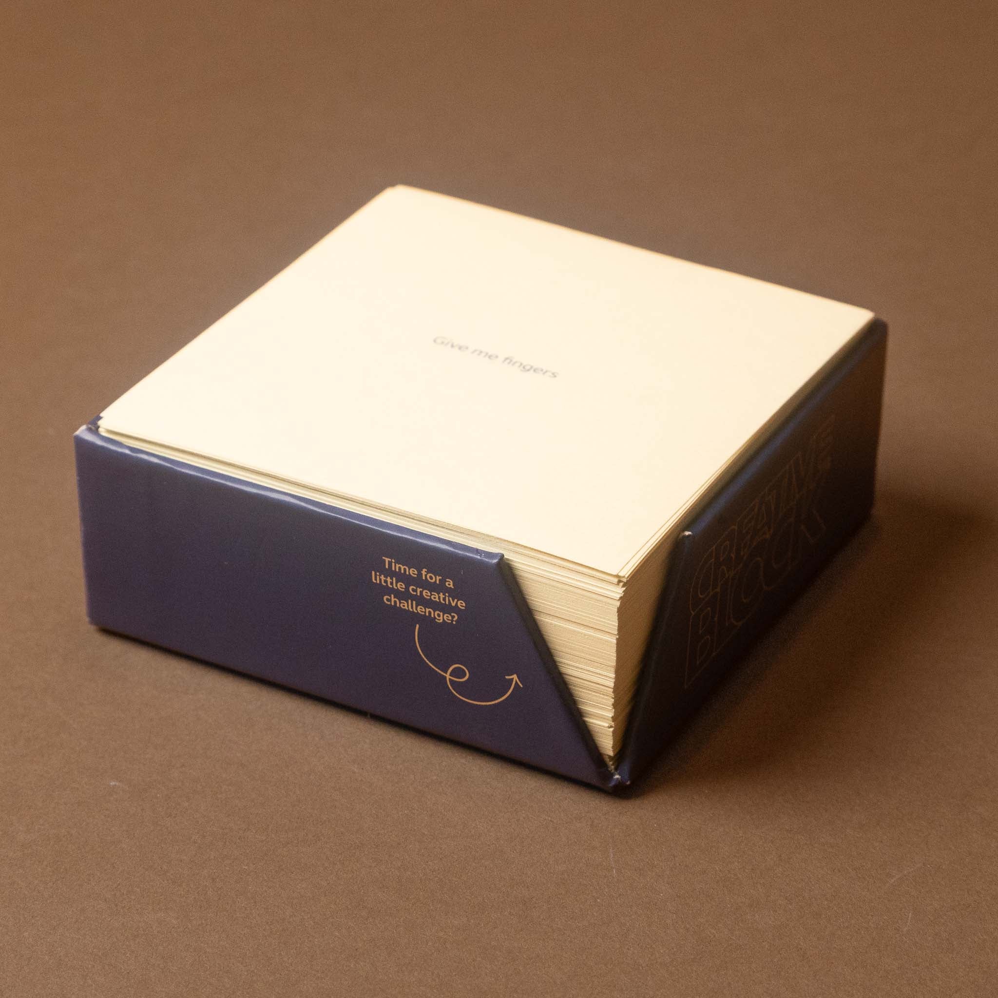 A boxed set of cream-colored cards or papers in a dark navy blue sleeve, photographed on a plain brown surface. The sleeve has gold text reading 'Time for a little creative challenge?' and a small gold arrow graphic.