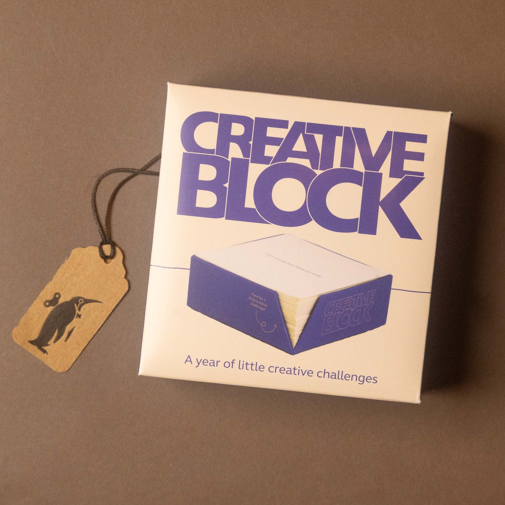 A square product box labeled 'CREATIVE BLOCK' with a hanging kraft paper tag printed with a penguin silhouette, photographed on a plain taupe-brown background.
