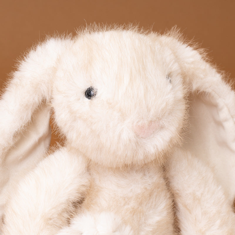 A close-up of a fluffy cream-colored plush bunny toy with long ears, black bead eyes, and a soft pink nose, set against a warm brown background.