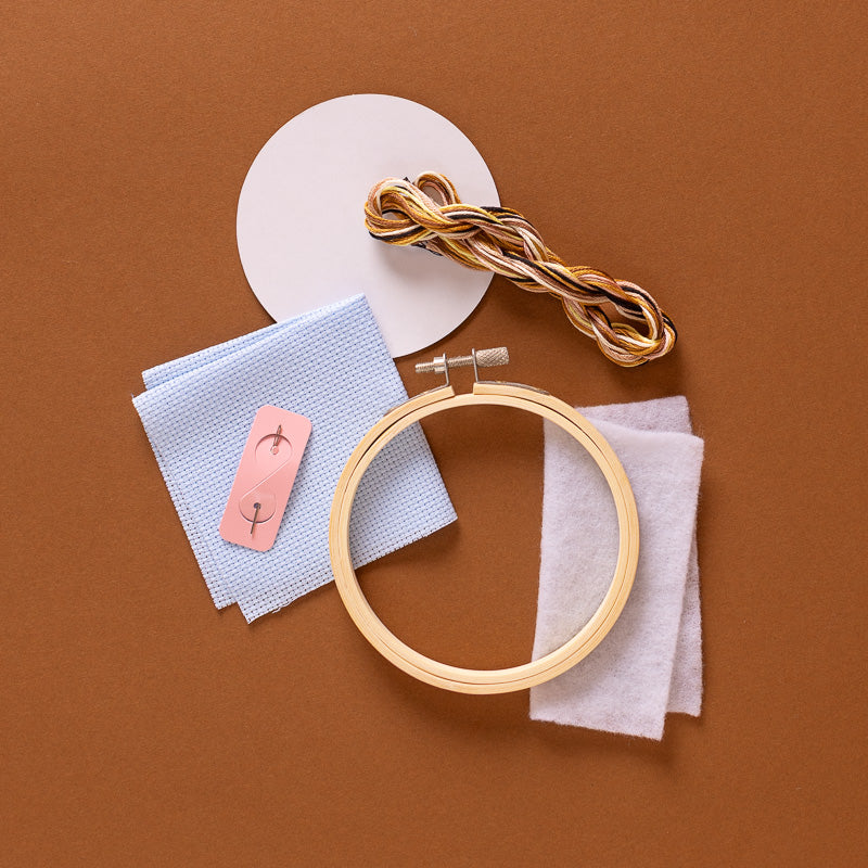 Embroidery kit components arranged on a brown background, including a wooden embroidery hoop, multicolor embroidery thread, a white circular template, light blue and white fabric pieces, and a pink sample patch with stitched design.