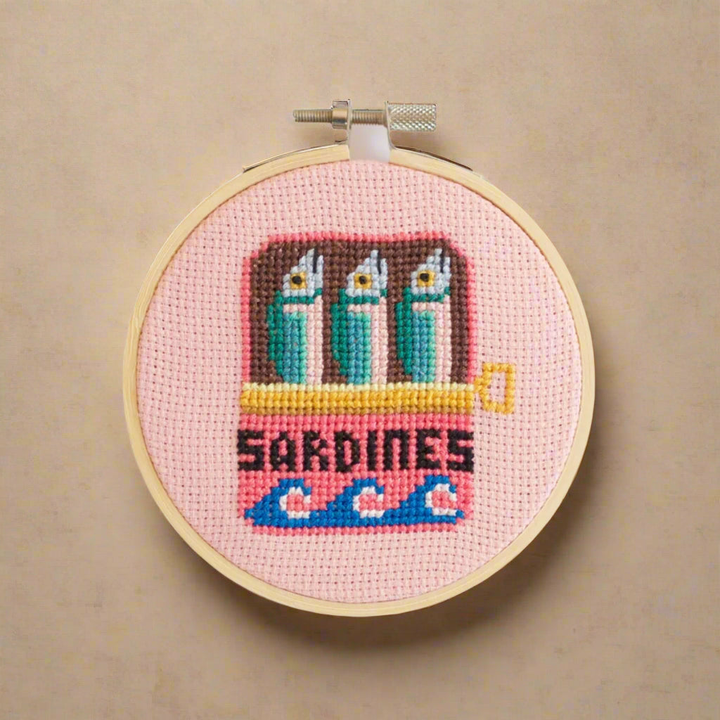 cross-stitch-kit-sardines in a can on a pink background, framed in a wooden hoop.