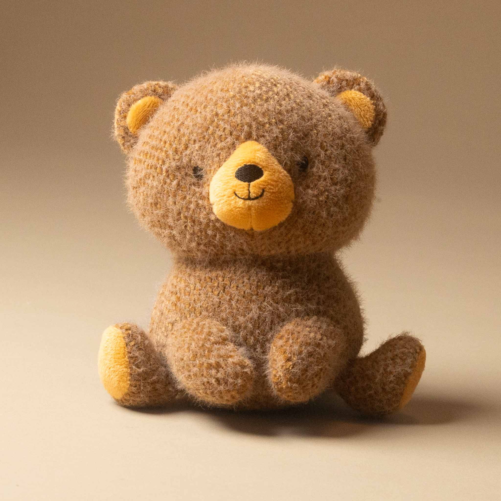 A soft, fuzzy cubby-bear-small plush toy in warm brown with a yellow snout, inner ears, and paw pads, sitting against a plain beige studio background.