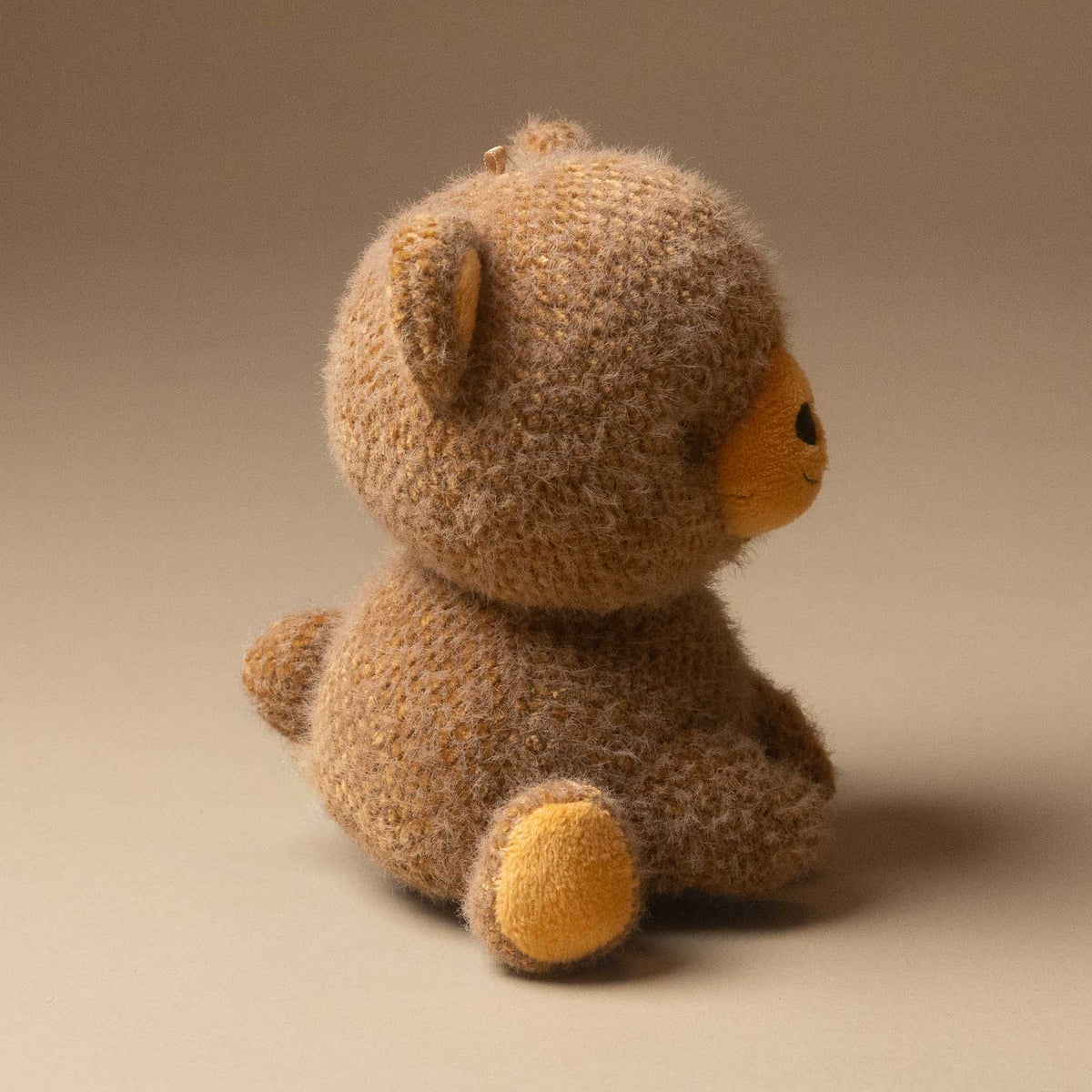 A cubby-bear-small plush toy shown from the back and side, with a warm brown body, mustard yellow snout, and yellow paw pad, photographed on a plain beige studio background.