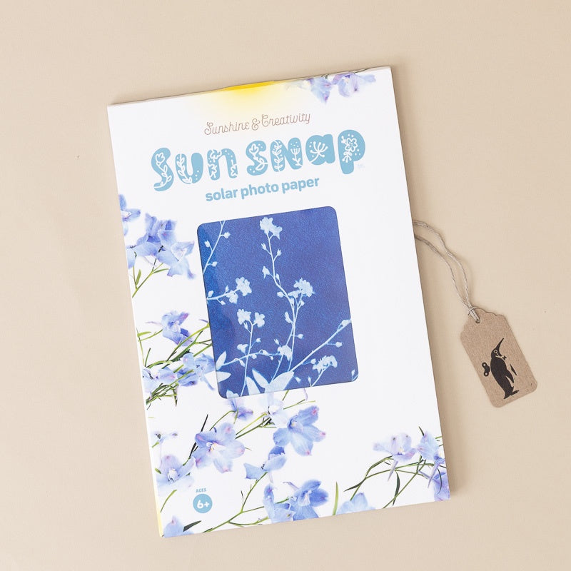 A package of Sunshine & Creativity Sunsnap Solar Photo Paper, featuring a white cover decorated with blue flowers and a blue sample photo area, shown on a beige background with a small paper tag attached.