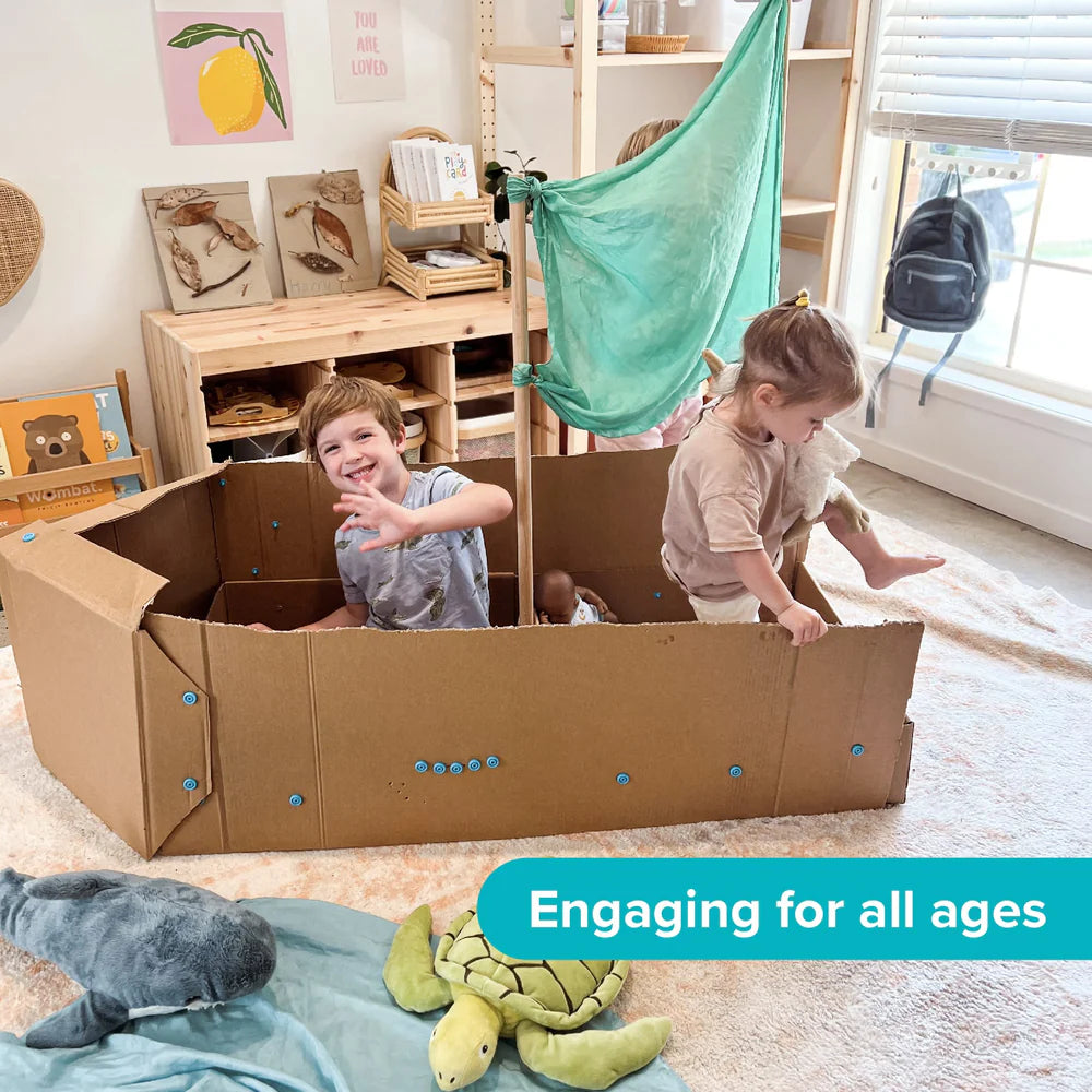 Two children playing with a cardboard boat in a room with toys and furniture.