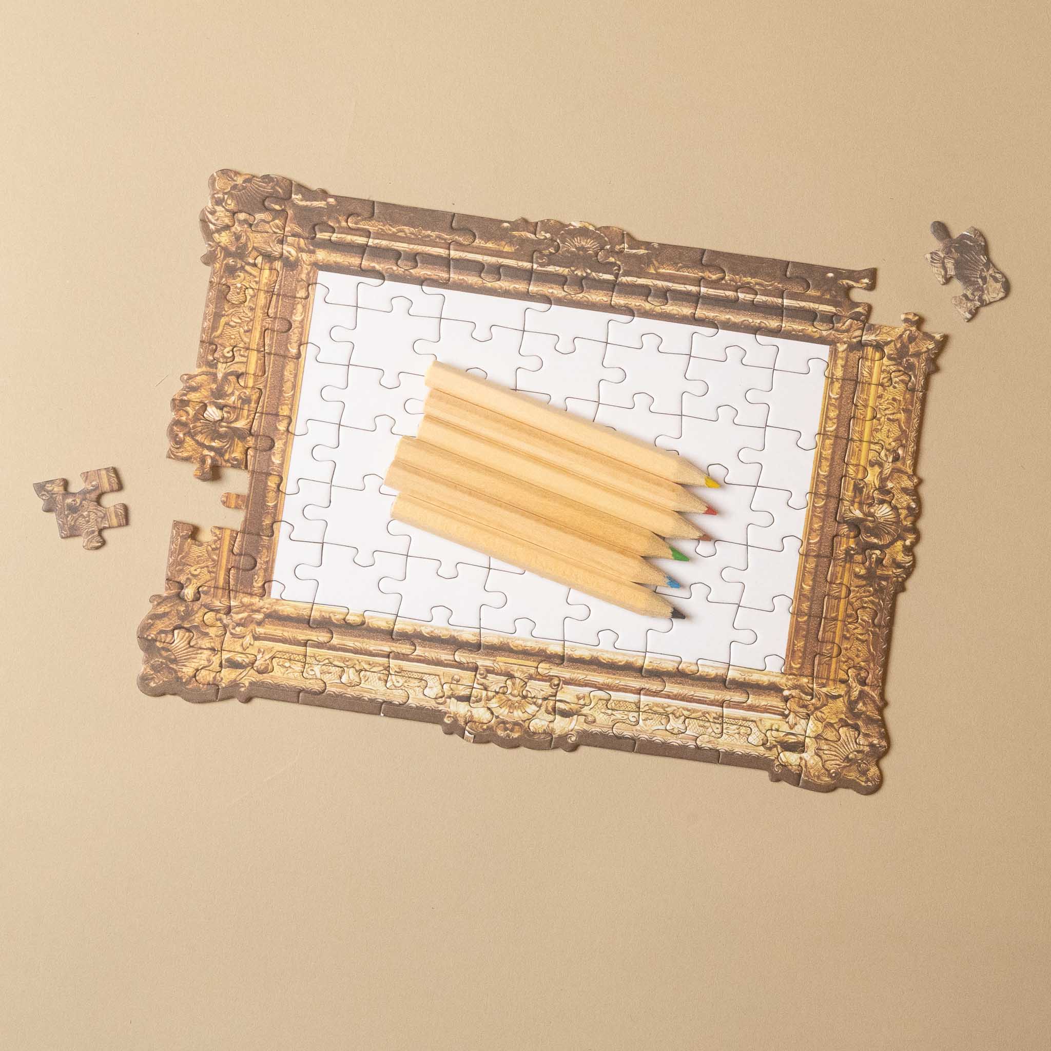 An ornate gold picture frame with a partially completed white jigsaw puzzle inside, topped with a stack of wooden pencils. A few loose puzzle pieces are scattered around on a warm beige background.