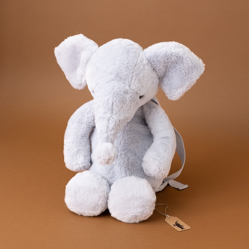 A light gray plush elephant toy with a small gray fabric backpack and a hanging paper tag, photographed against a solid warm brown studio background.