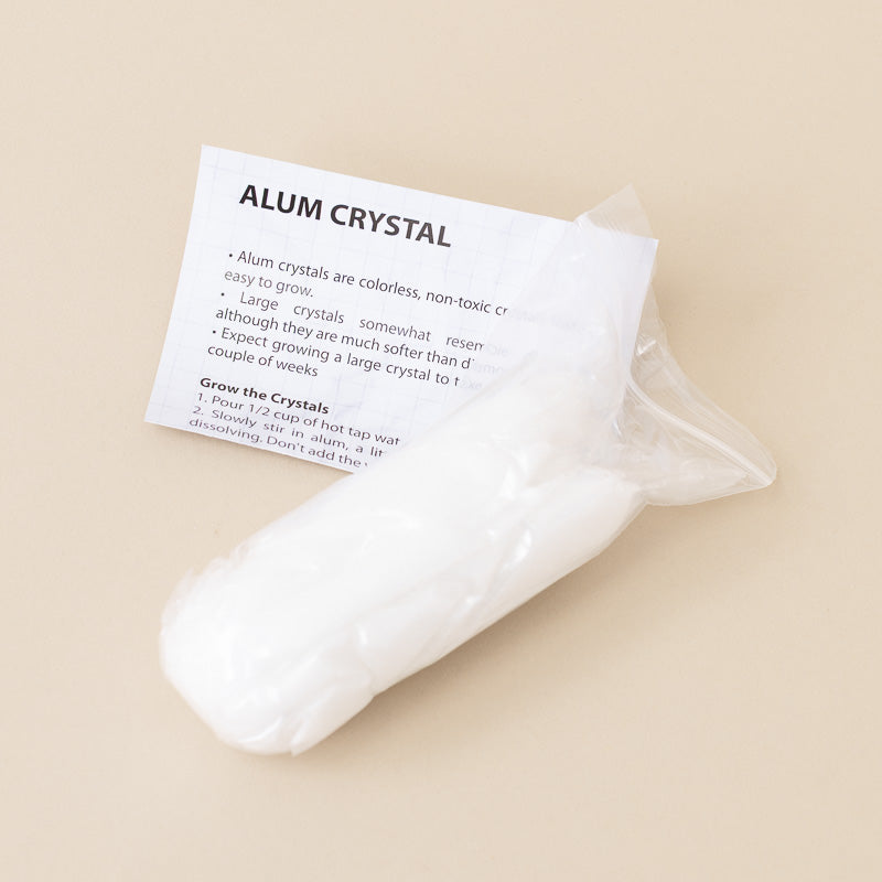 A clear plastic bag containing white alum crystals with an attached printed card that reads 'ALUM CRYSTAL' and includes growing instructions, photographed on a warm beige background.
