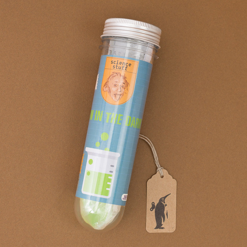 A clear plastic tube with a white screw cap and a blue label reading 'science stuff' and glow in the dark containing a green object at the bottom, photographed on a brown background with a small hanging tag attached.