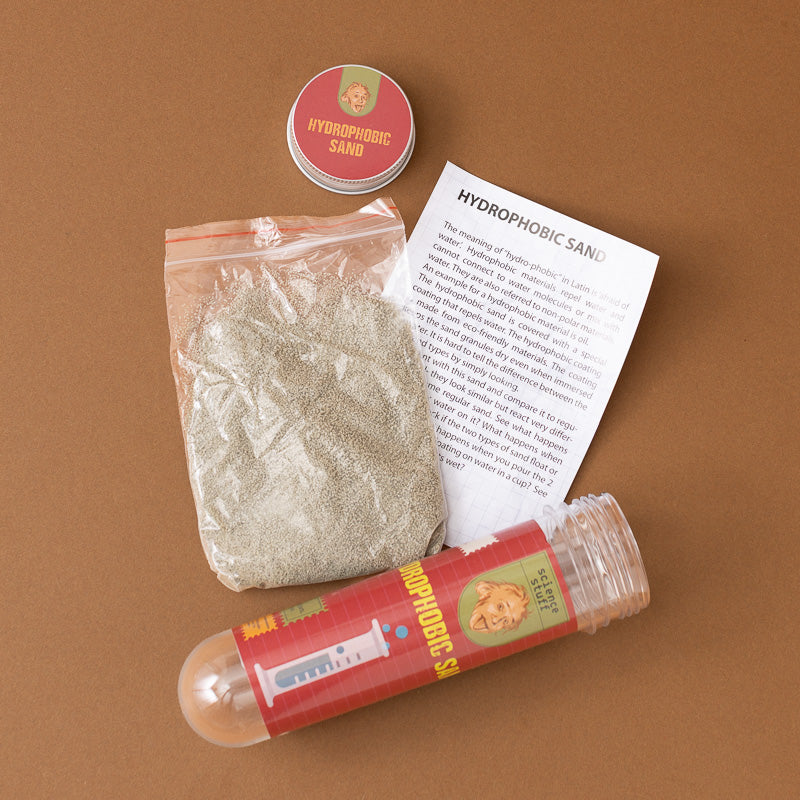 Hydrophobic sand kit with a clear plastic bag of light gray sand, a small round tin labeled 'HYDROPHOBIC SAND', an instruction sheet, and a clear cylindrical tube with a red label, arranged on a brown background.