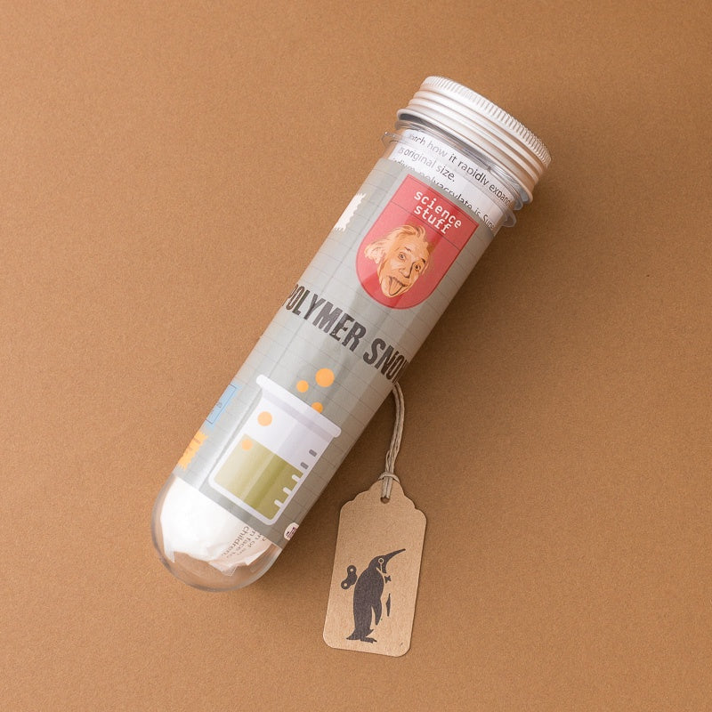 A clear plastic tube containing white powder, labeled 'science story' and 'POLYMER SNOW', with a small paper hangtag featuring a black penguin silhouette, set against a solid brown background.