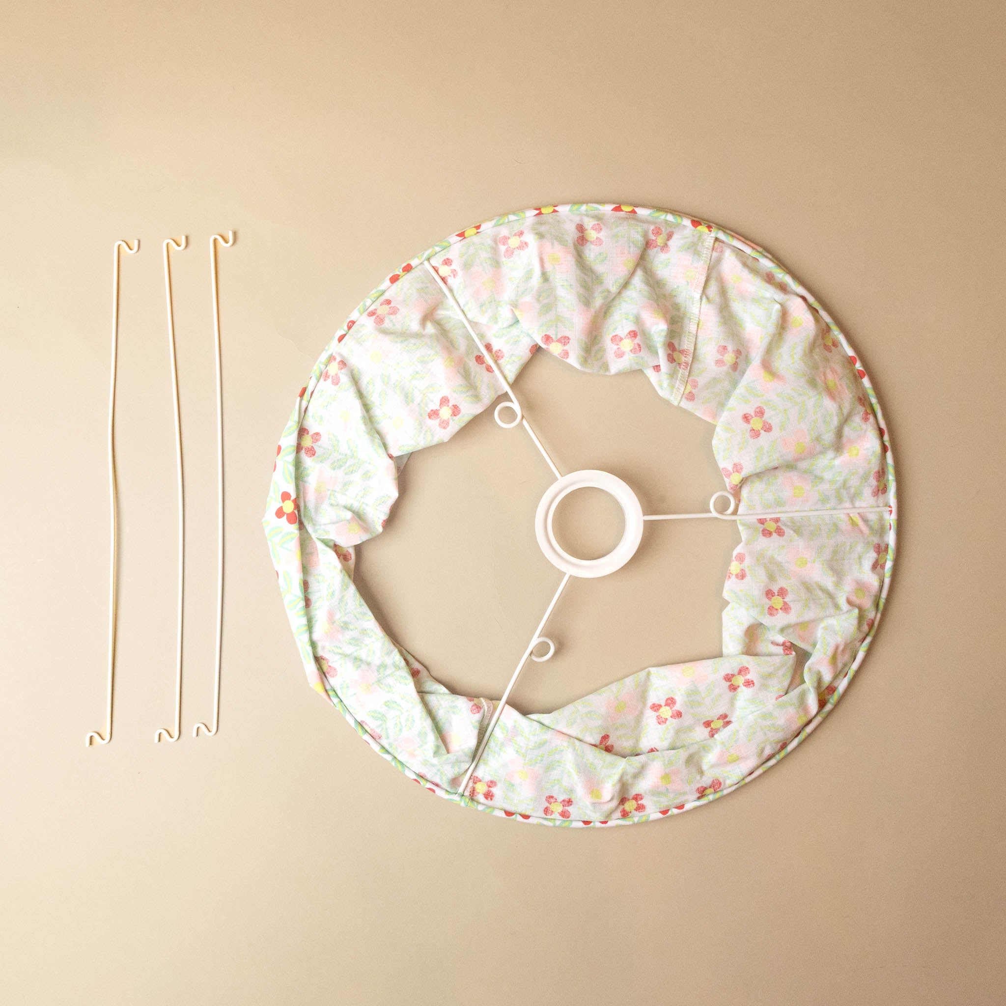fabric-lantern-bindweed_ A circular fabric shade or skirt with a small floral print in pink, orange, and green on a white base, shown laid flat on a beige background alongside three long metal support wires.