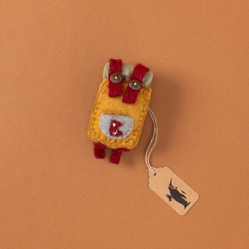 A small handmade felt backpack in mustard yellow, red, and light gray, with two metal buttons on top and a paper tag hanging from a string, photographed on a solid warm brown background.