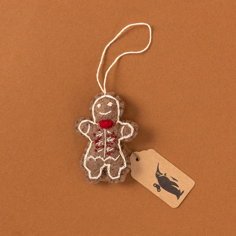 A gingerbread man felt ornament with white embroidered outlines, a red bow tie, and a small red floral chest detail, hanging from a white string on a brown background with a kraft paper tag attached.