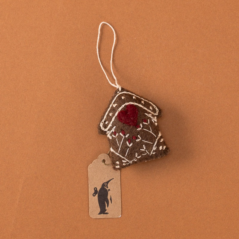 A small brown felt ornament house with white embroidered details, a red pom-pom, and a white hanging string, shown on a warm brown background with a small kraft paper tag featuring a black penguin silhouette.
