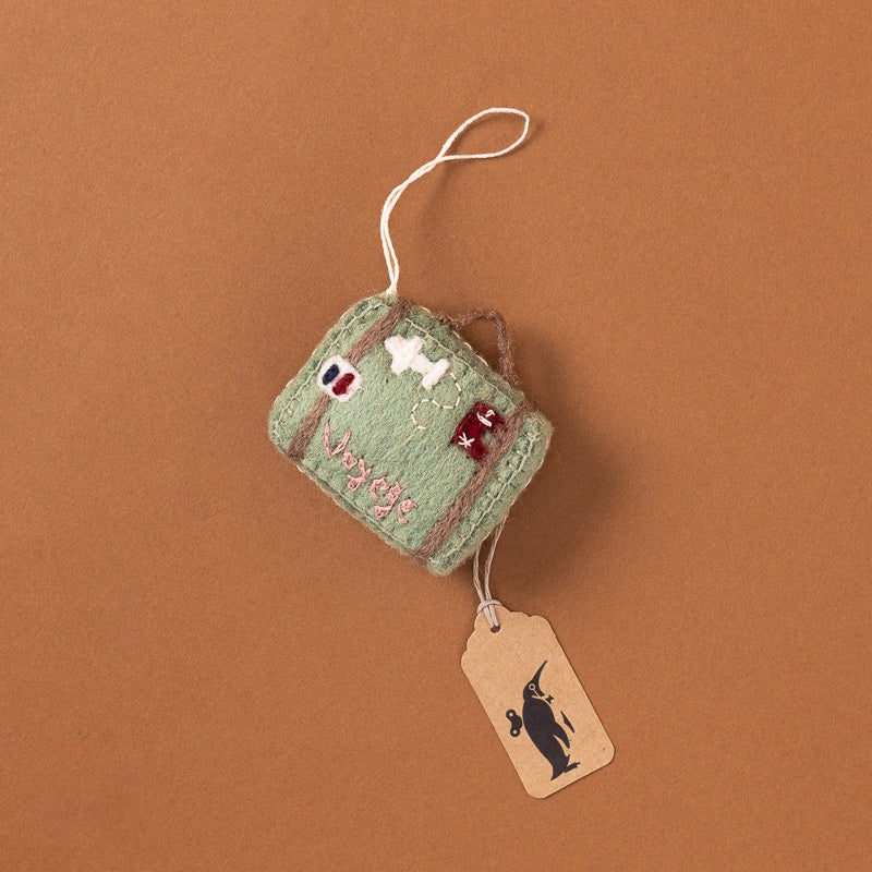 A small handmade ffelt-embroidered-suitcase-ornament-sage with embroidered details, shown on a warm brown background with a paper tag attached.