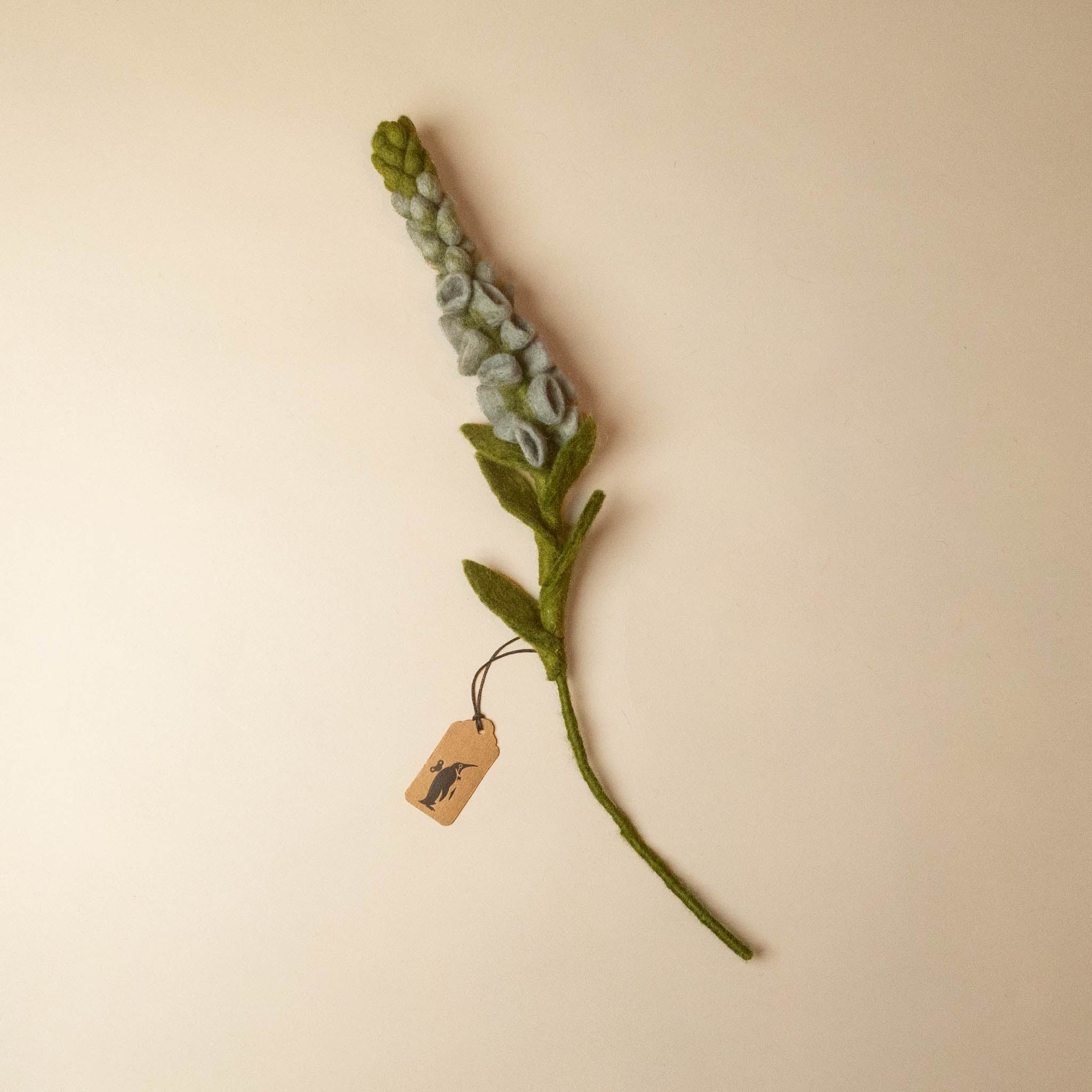 felt-foxglove-flower-beau-blue with pale blue-green petals and green leaves, attached to a brown paper tag, photographed on a plain warm beige background.