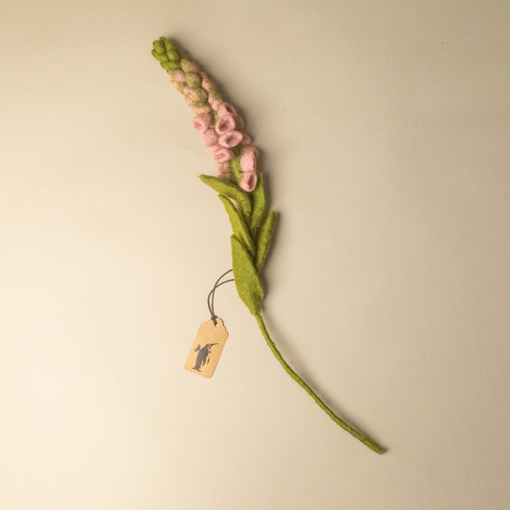 A handcrafted felt-foxglove-flower-blushfeaturing light pink blossoms, green leaves, and a long green stem, with a small paper tag attached, set against a plain beige background.