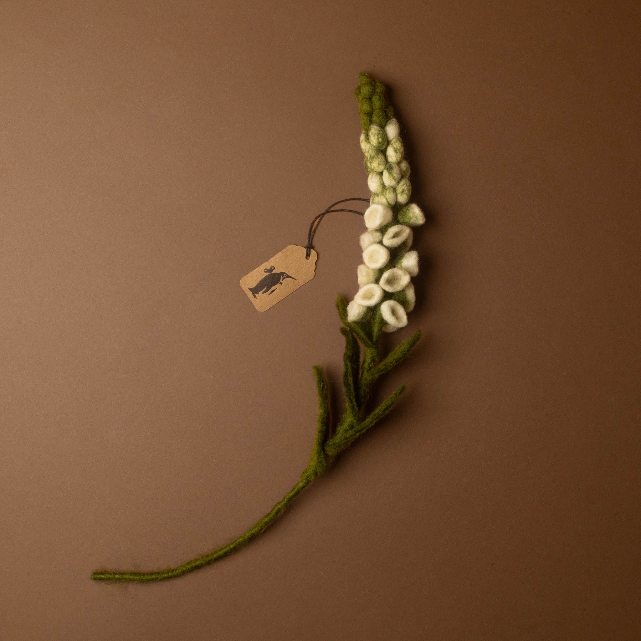 A handcrafted felt flower stem featuring white felted blossoms and green felted leaves and stem, with a small paper tag attached, set against a solid brown background.