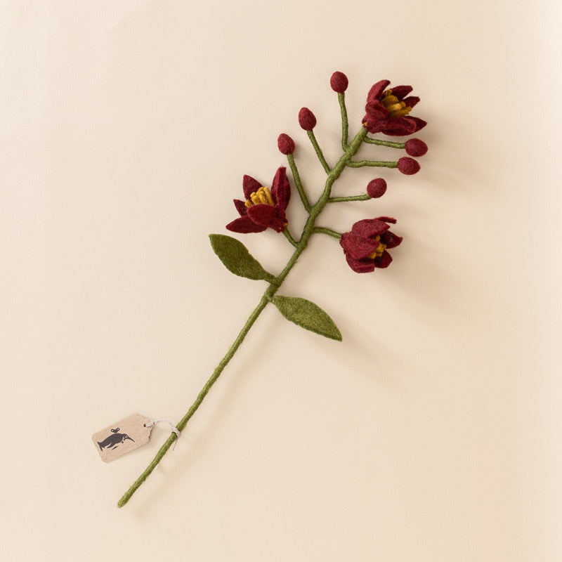 A single artificial flower stem with dark red felt blossoms, green leaves, and a green stem, photographed on a plain warm beige background. A small paper tag is attached near the bottom.