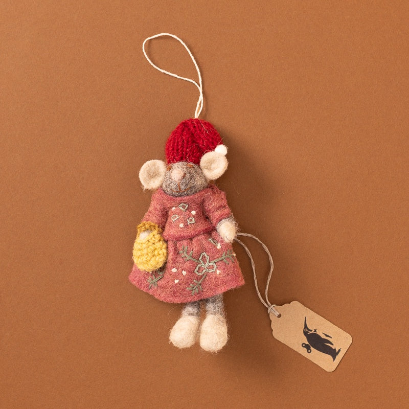 A felted wool mouse ornament wearing a red knit hat and pink dress with embroidered floral details, holding a yellow crocheted basket, shown on a warm brown background with a hanging paper tag.