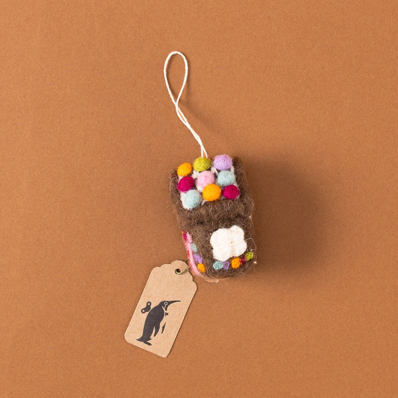 A small felted wool house, with a white hanging loop and a kraft paper tag printed with a penguin silhouette, set against a solid brown background.