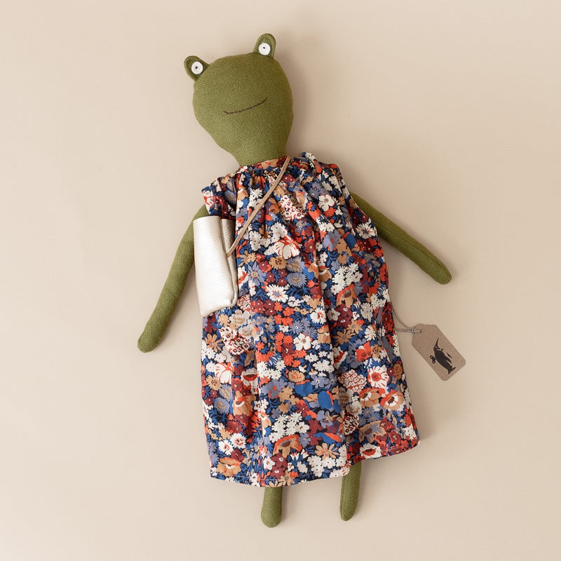A green plush frog doll wearing a sleeveless floral dress with a blue, red, orange, and white pattern, posed against a solid beige background with a small white bag and a hangtag.