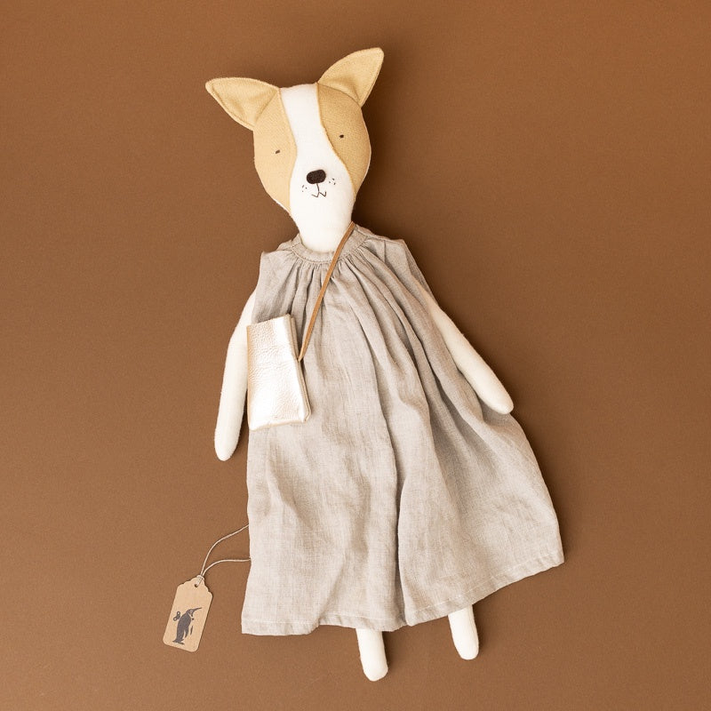 A handmade plush dog doll with a tan and white face, wearing a light beige linen dress and holding a small matching pouch, set against a solid brown background.